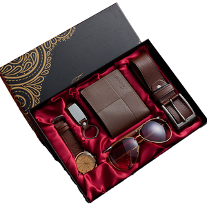 Open black and gold gift box on red satin holding a brown leather wallet, a leather belt, a watch, sunglasses and a keyring